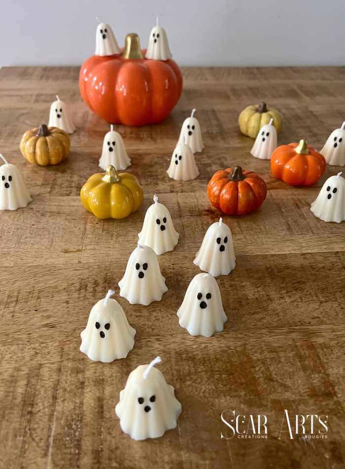 Bougies en forme de petits fantômes blancs disposées en file sur une table en bois, entourées de citrouilles jaunes et orange.

