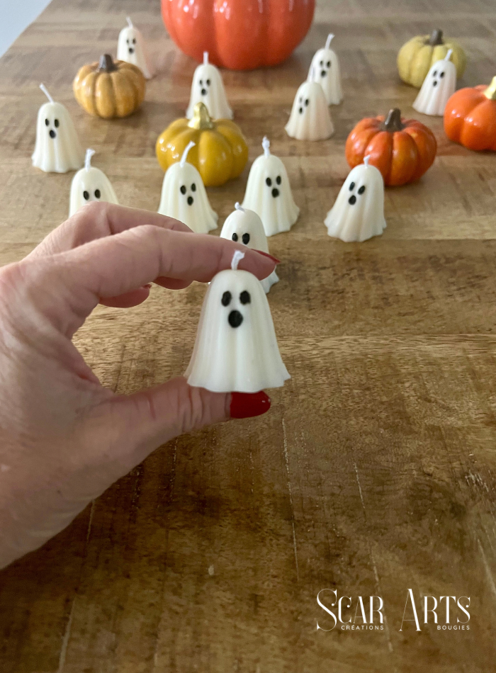 Petite bougie fantôme blanche tenue entre les doigts, devant un ensemble de fantômes blancs et de citrouilles sur une table en bois.