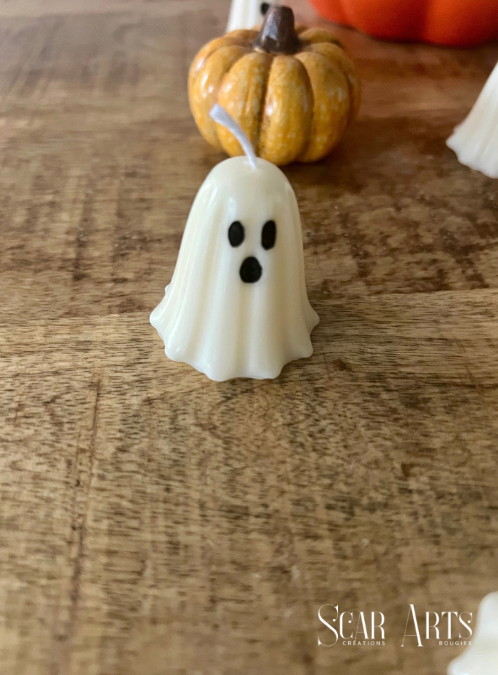 Petit fantôme blanc en bougie, vu de face sur une table en bois, avec une citrouille jaune à tige brune en arrière-plan.

